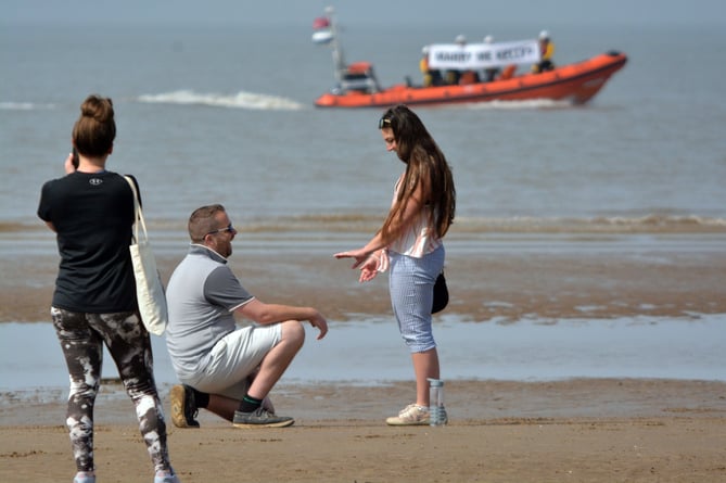 Lyndon pops the question. Photo released May 15 2024. RNLI volunteer surprises partner with unique proposal - with lifeboat crew holding up huge banner on the waves.Lyndon and Kelly were walking along Burnham-On-Sea beach on Sunday (12 May) - when a lifeboat appeared on the waves with RNLI crew holding a three metre banner stating: Marry me, Kelly?.After nearly 20 years together, Lyndon, the Burnham-on-Sea RNLI Deputy Press Officer, decided there was no better place to get down on one knee than on the coastline in front of RNLI's Lower Lighthouse.Lyndon, who had spent spent weeks planning the very special moment with the help of crew, said: "Thankfully she said yes!"
